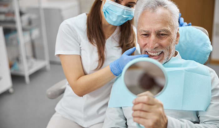 Older man sitting in a dental chair looking in a handheld mirror at his new dental implants from David Paulussen, DMD in Hackettstown, NJ